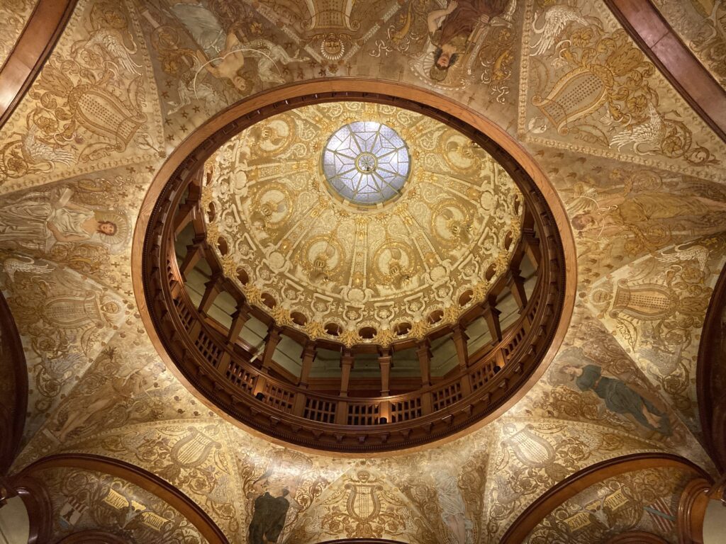 A gold patterned dome with a glass window in the middle and a beautiful mural painted ceiling